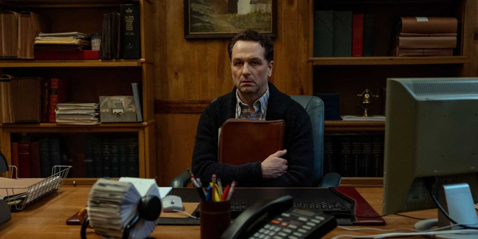Matthew Rhys como Prefeito Tom Loftis em Widow's Bay, segurando um livro e com expressão nervosa.