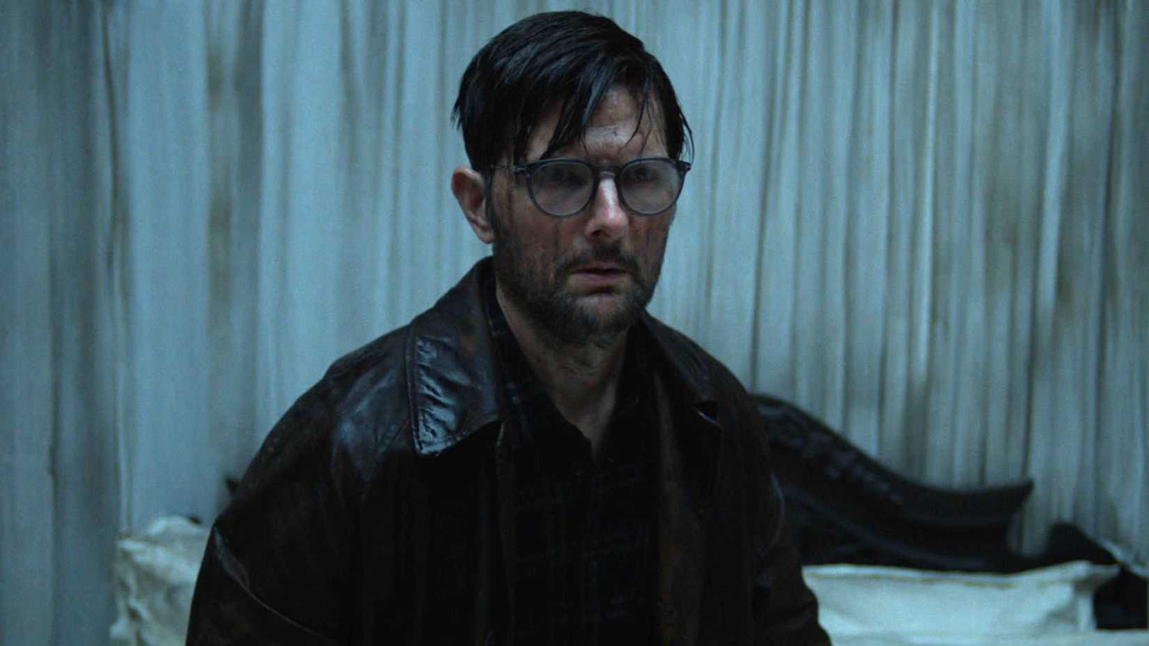 Adam Scott covered in blood and holding a lantern in a curtained room in Hokum