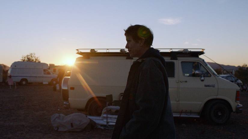 Fern de perfil caminhando por uma van ao pôr do sol em Nomadland