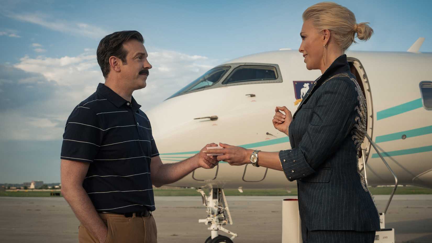 Ted Lasso (Jason Sudeikis) entrega uma caixa de biscoitos para Rebecca Welton (Hannah Waddingham) perto de um jato particular na 4ª temporada de Ted Lasso.