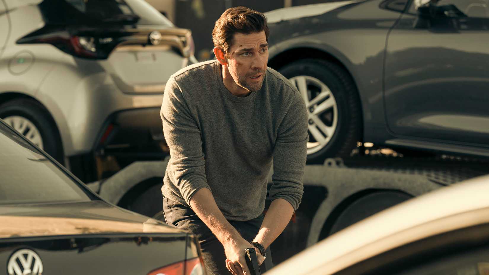 John Krasinski como Jack Ryan correndo por um grupo de carros com a arma em punho na 4ª temporada de Jack Ryan.