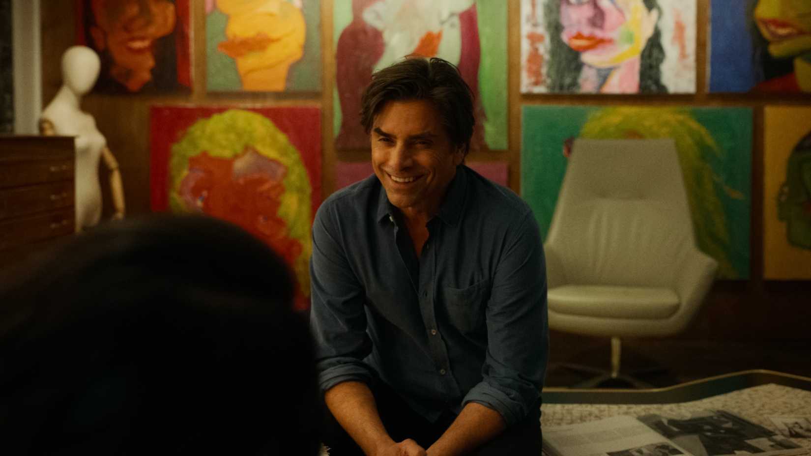 john stamos sits in front of paintins and smiles in the drag