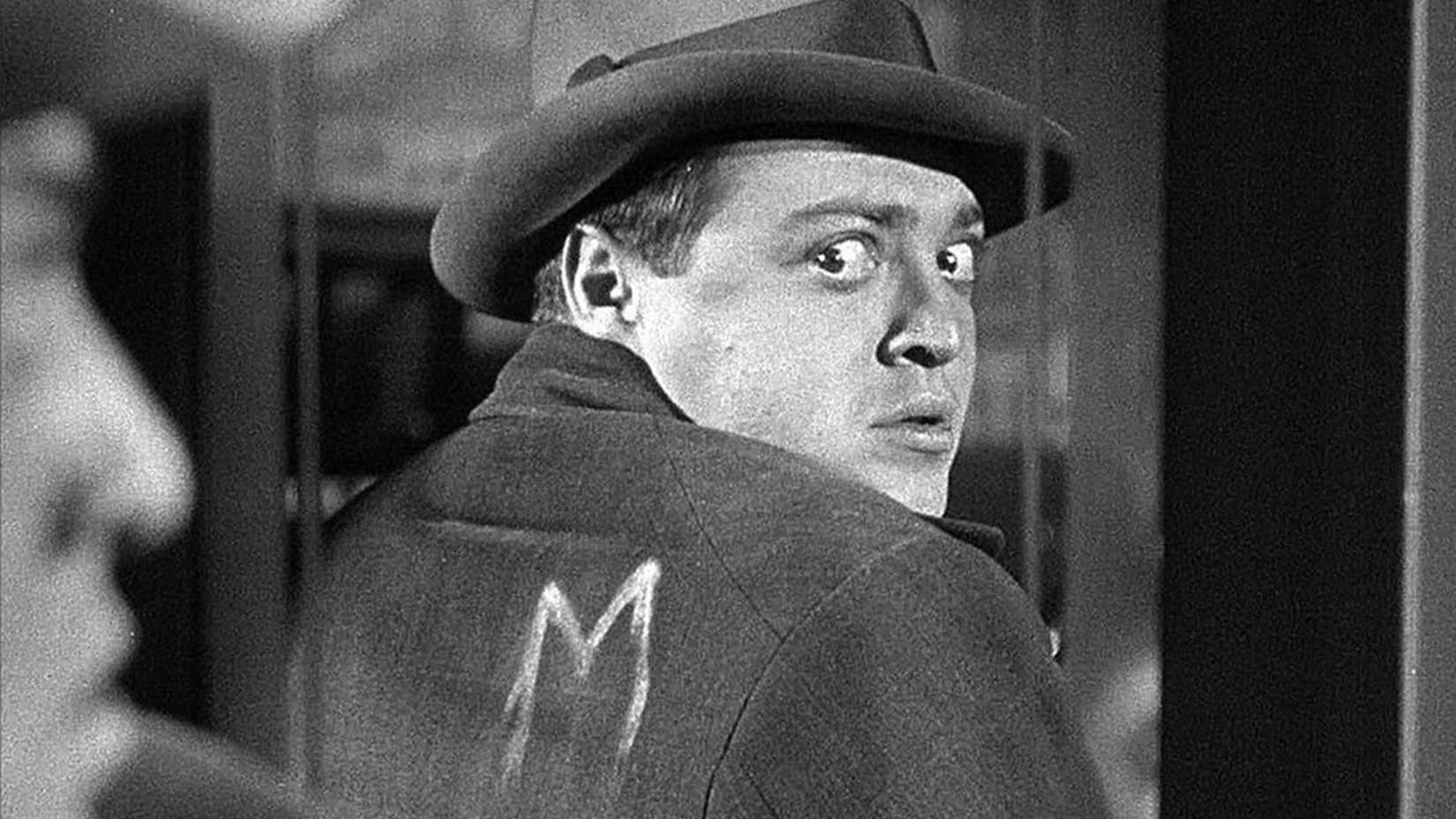 Peter Lorre olha por cima do ombro para ver a letra M em seu casaco em M
