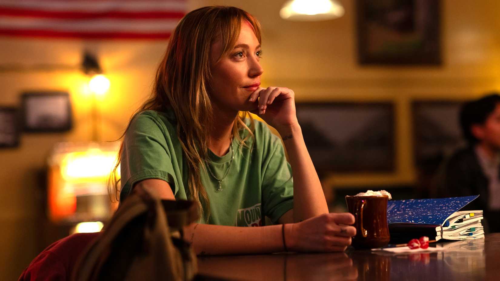 Maika Monroe's Kenna looking in through while sitting at a bar with a hot chocolate in Reminders of Him