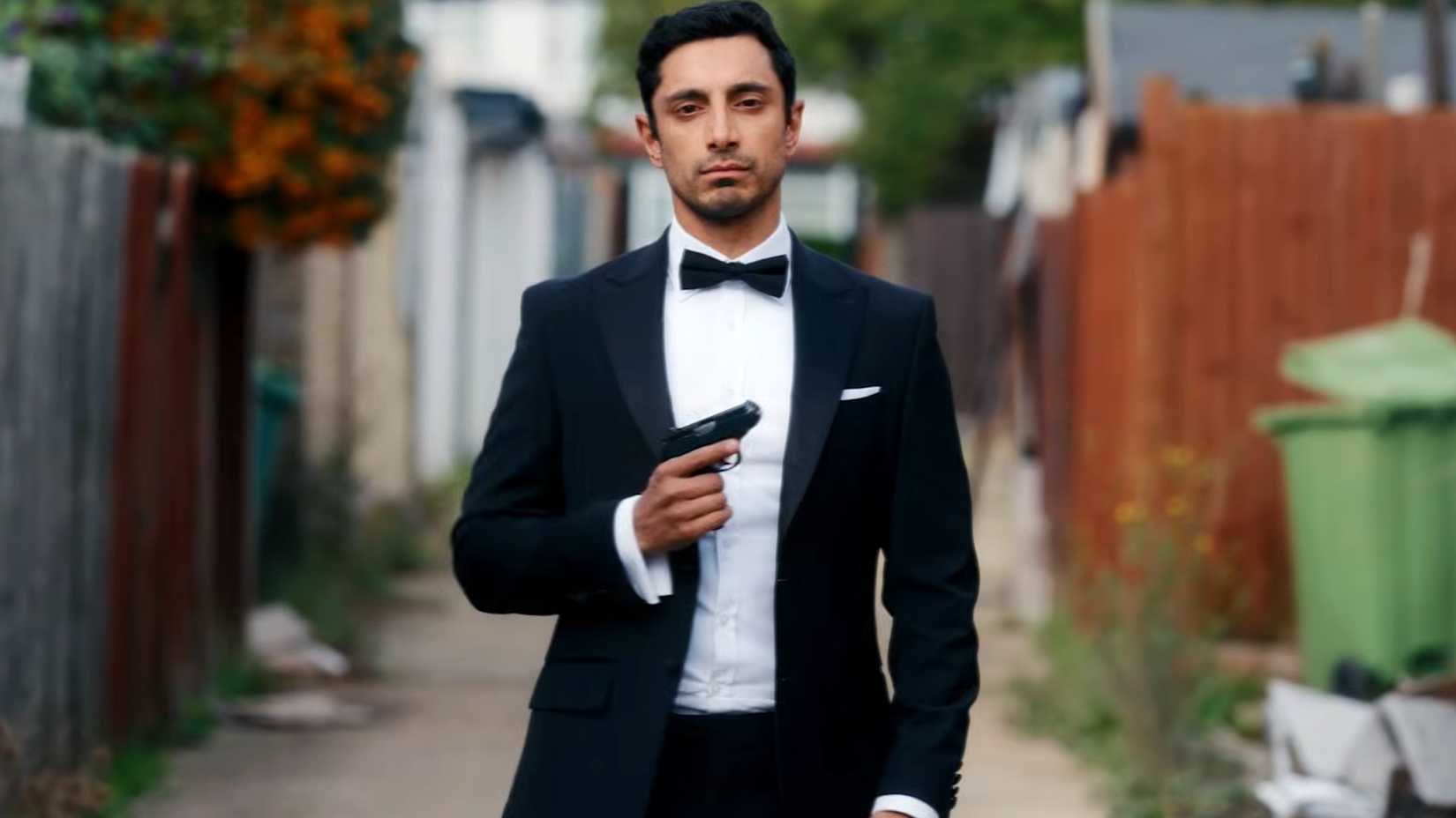 Riz Ahmed em smoking com pose de arma na série Bait.