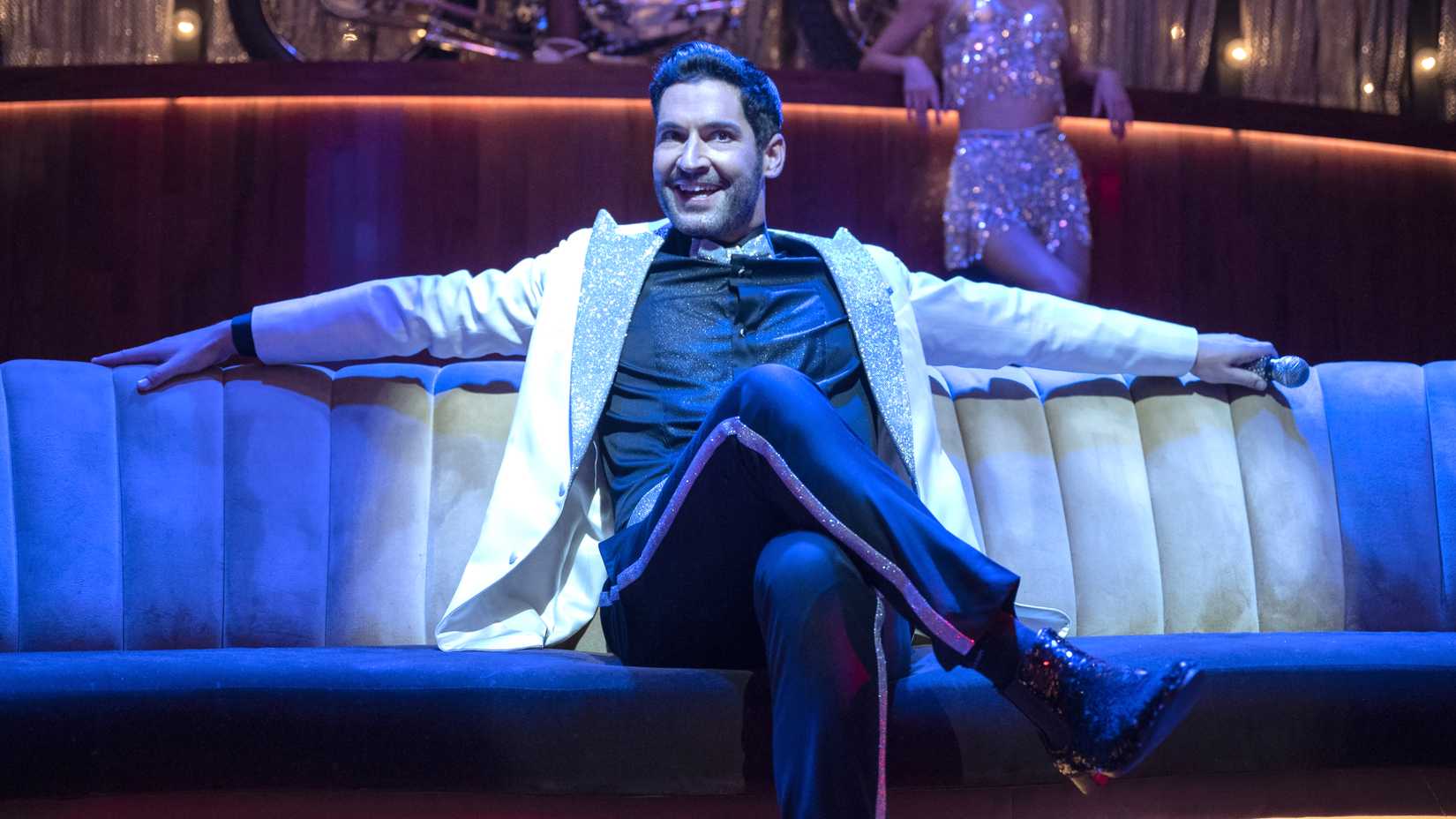 Tom Ellis' Lucifer sitting on a couch with a spotlight on him for a performance in Lucifer season 6