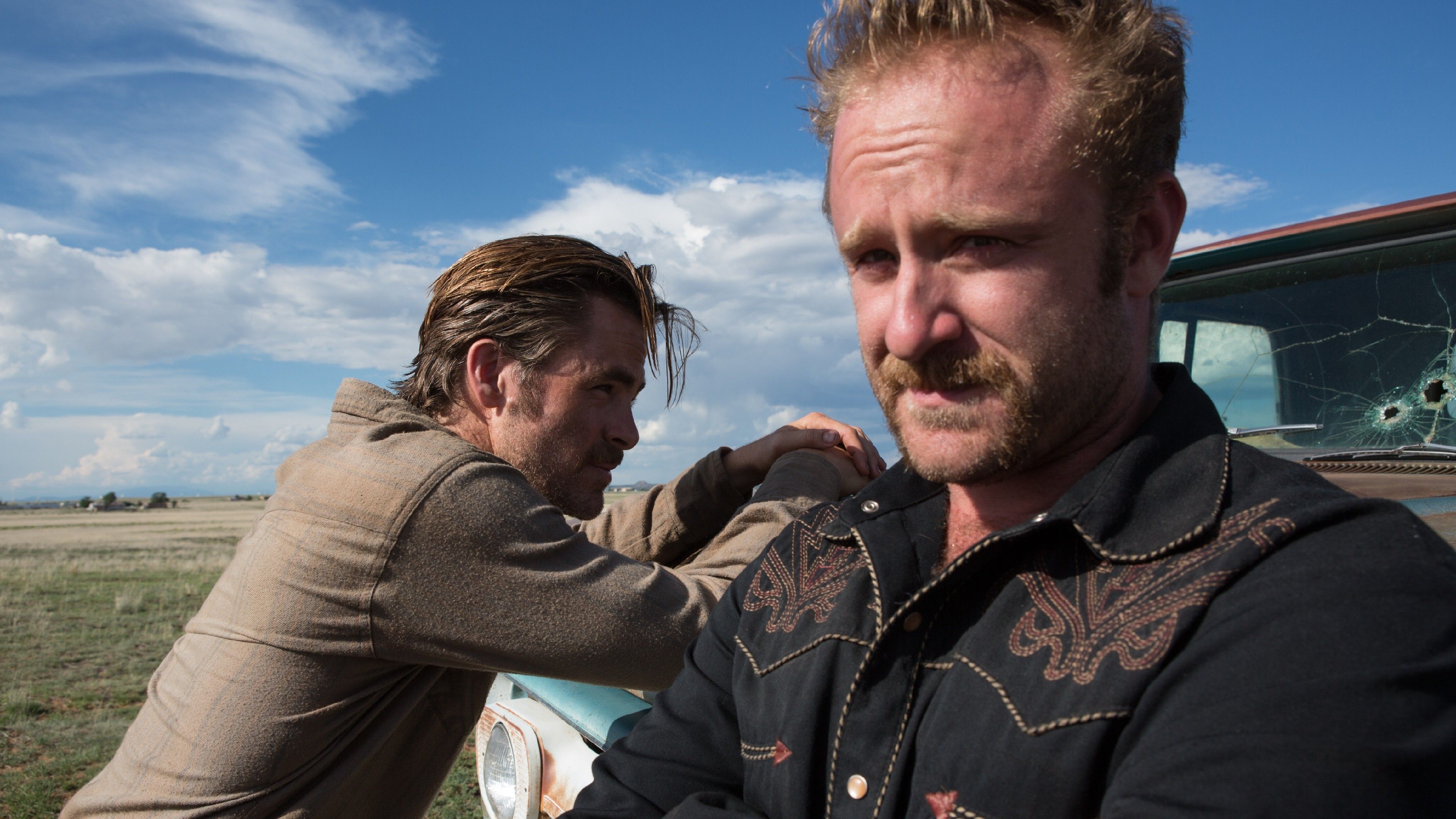 Chris Pine leans toward the hood of a truck with bullet holes in it and Ben Foster faces the other direction and looks into the distance in Hell or High Water