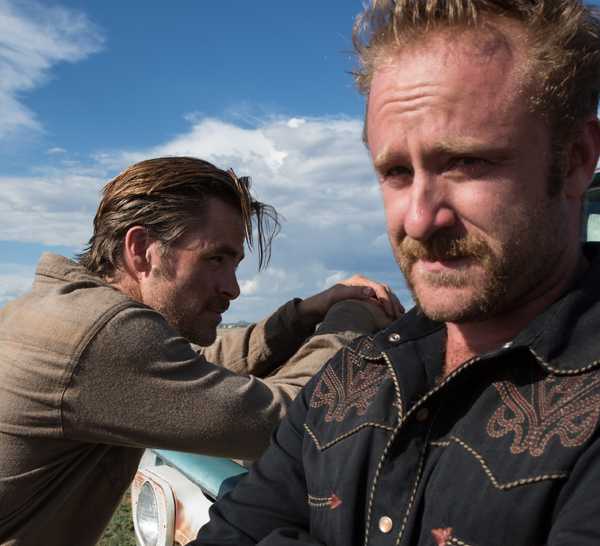 Chris Pine leans toward the hood of a truck with bullet holes in it and Ben Foster faces the other direction and looks into the distance in Hell or High Water