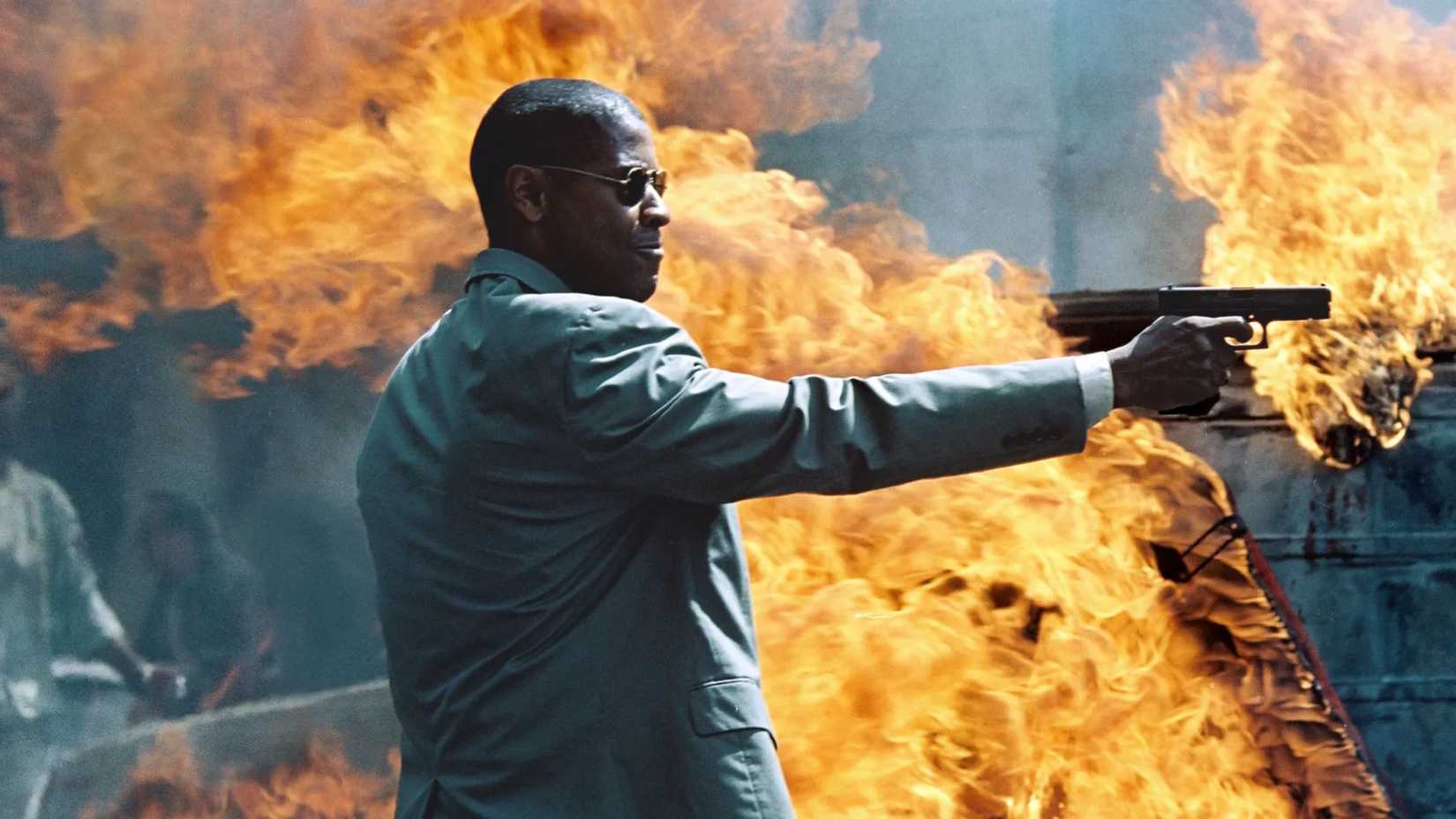 Denzel Washington's John Creasy shoots a gun with a fire behind him in Man on Fire