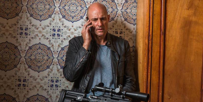 Mark Strong sentado com um rifle no colo enquanto fala ao telefone em Deep State