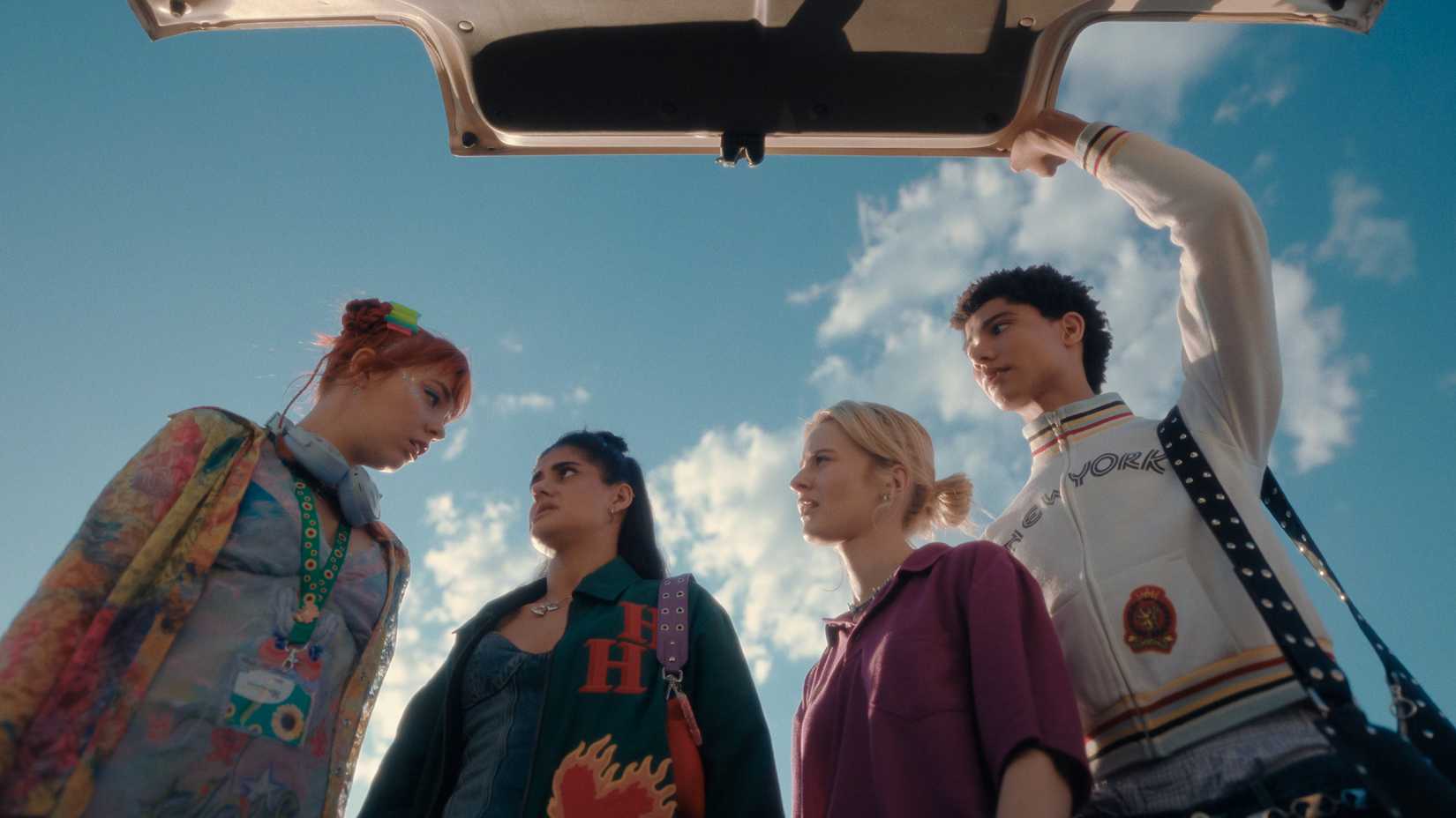 Quinni, Harper, Amerie, and Darren shot from underneath as they open a car trunk in Heartbreak High season 3. There's a blue sky above them.