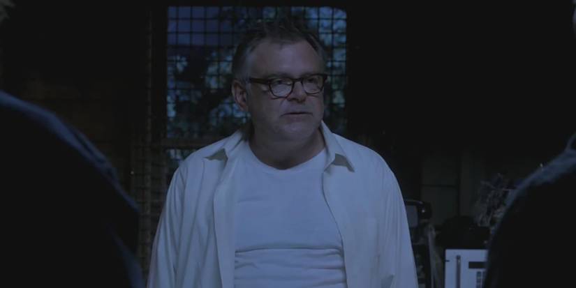 Still de Supernatural com Frank Devereaux em um quarto escuro usando uma camisa branca de botões, camiseta e óculos.