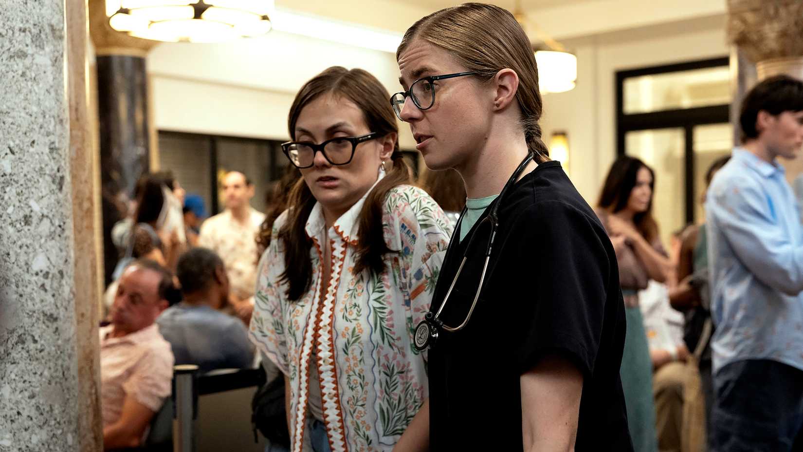 Tal Anderson como Becca e Taylor Dearden como Dr. Mel King, com uma expressão preocupada enquanto se movem pela sala de espera em The Pitt Temporada 2