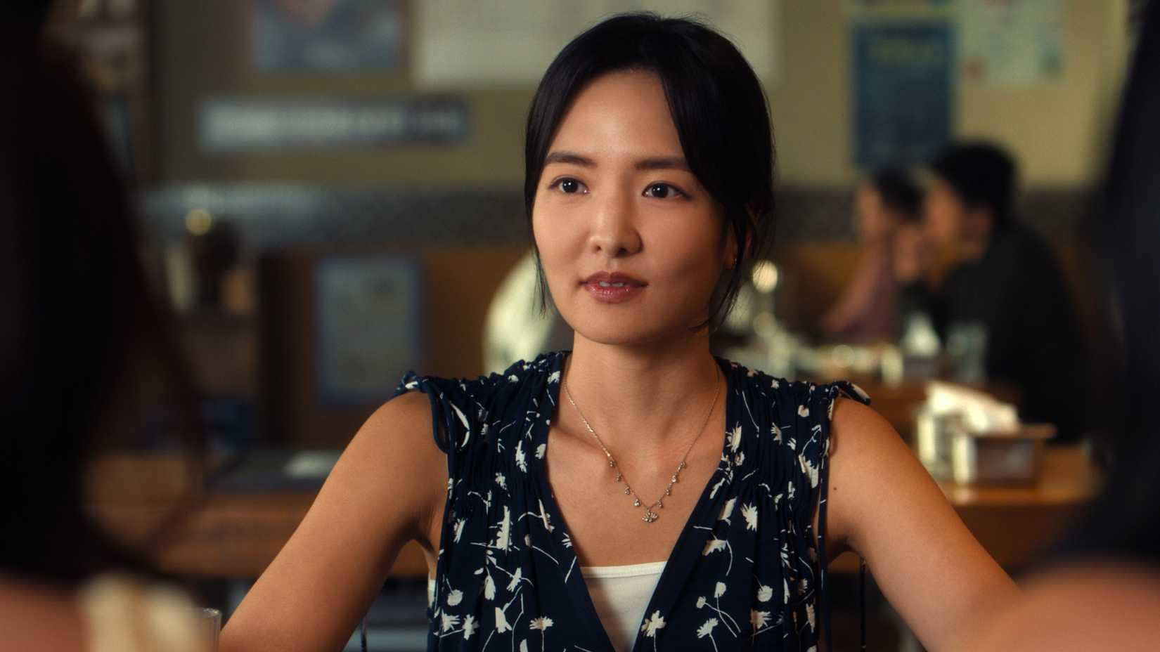 Kitty & Min Ho's Relationship Status 1 XO Kitty still featuring Jiwon Ahn sitting at a table across from Kitty and Lara Jean.