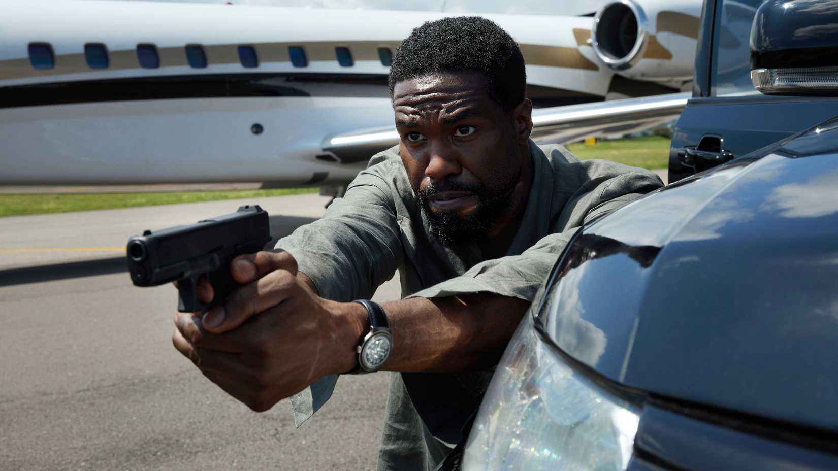 Yahya Abdul-Mateen II Creecy aims a gun while hiding behind a truck in front of a jet in Man on Fire
