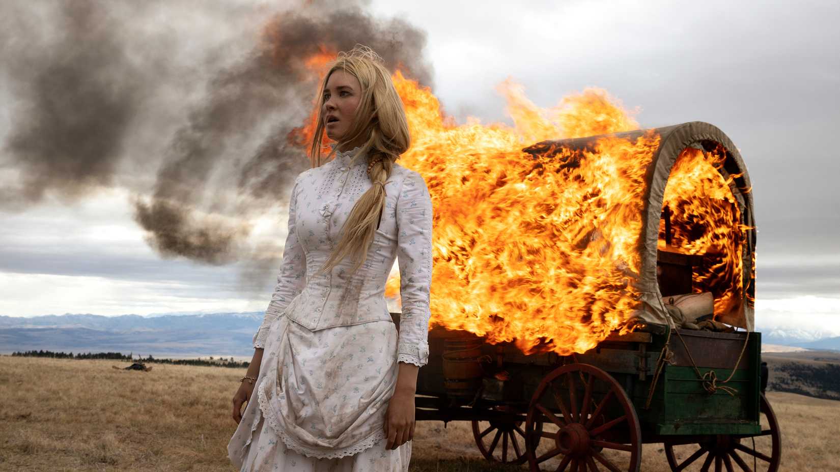 1883 still featuring Isabel May as Elsa Dutton, wearing a white dress and standing in front of a burning covered wagon.