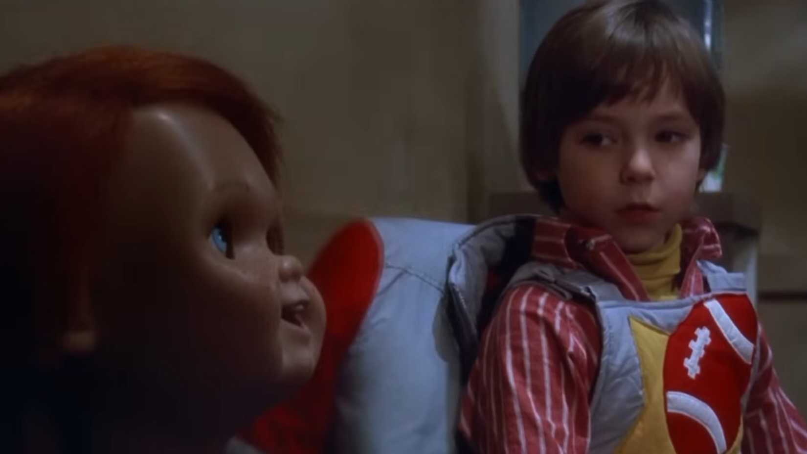 A kid looking concerned sitting next to a Chucky doll in Childs Play