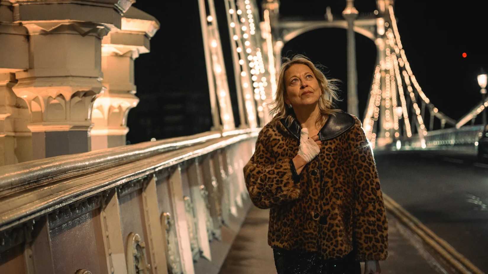 Alice and Steve still featuring Alice in a leopard print jacket with her hand bandaged, walking over a bridge.