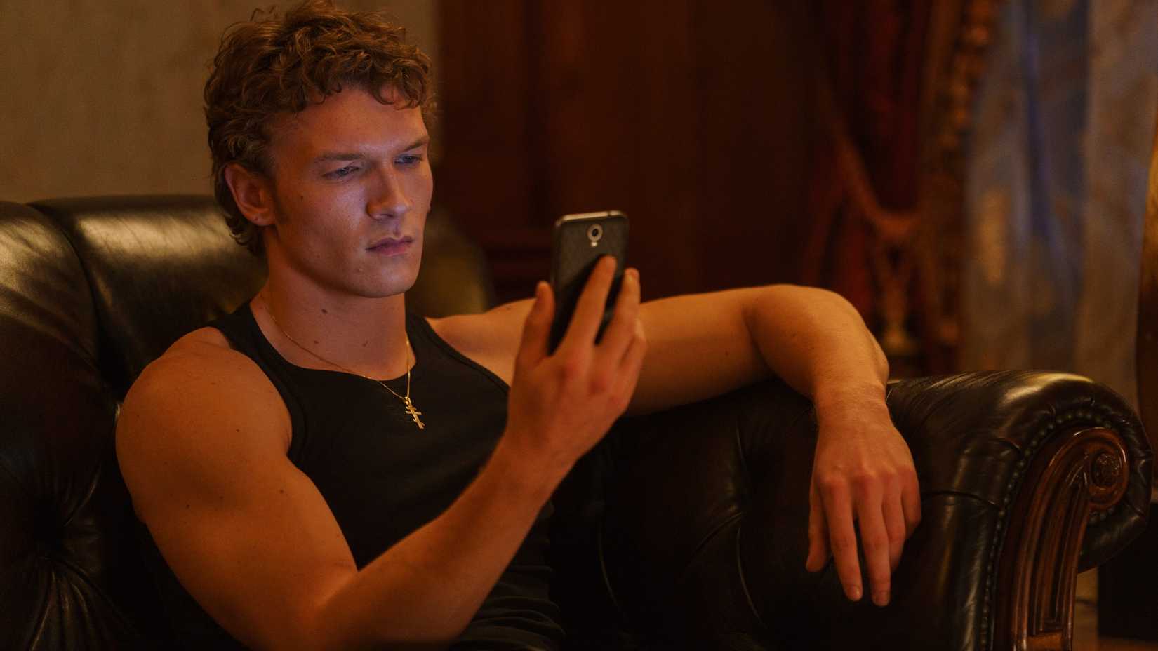 Heated rivalry of Connor Storrie as Ilya Rozanov sits on a leather couch with his phone.