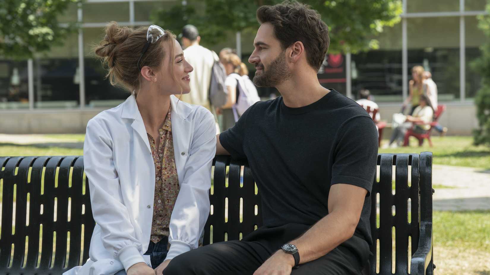 Lili Reinhart as Olive and Tom Bateman as Adam sitting on a bench and smiling at each other in The Love Hypothesis