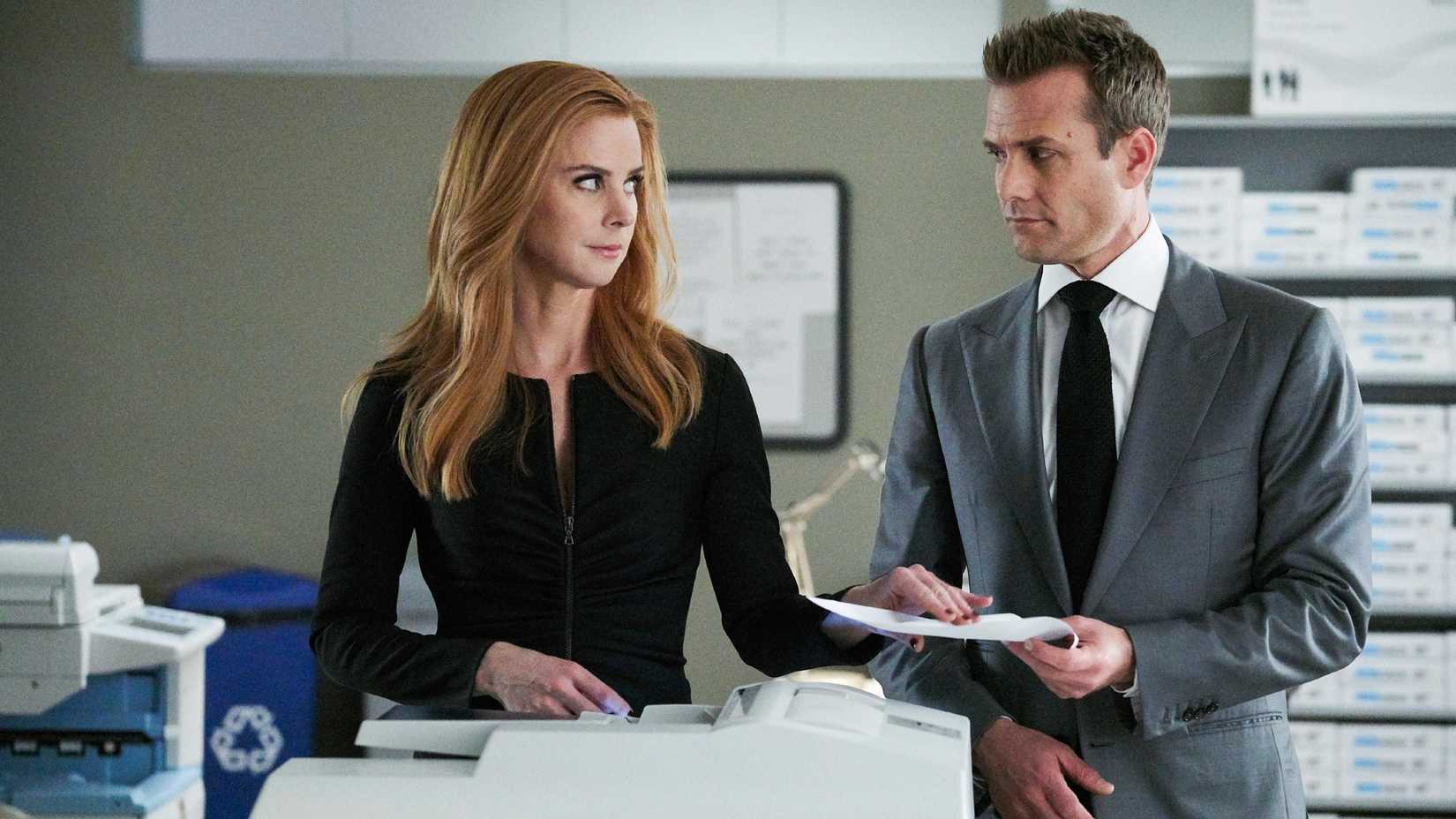 Suits still featuring Sarah Rafferty as Donna and Gabriel Macht as Harvey, standing over a copy machine and smirking at each other.