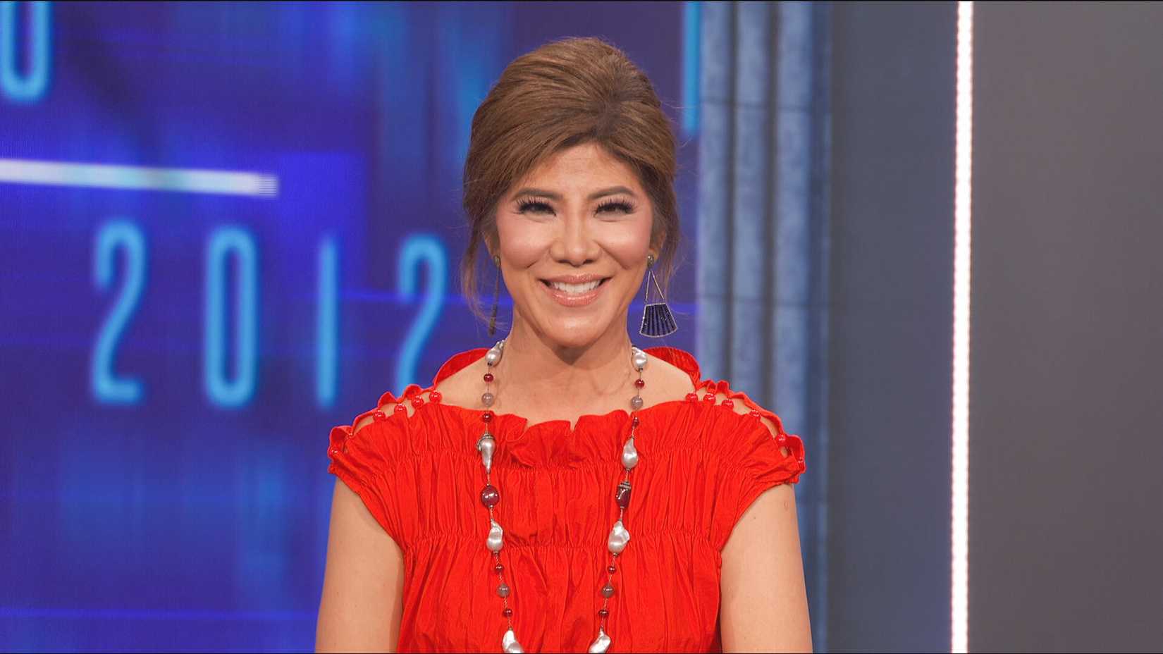 Still of Julie Chen Moonves on the Big Brother stage, smiling in a red dress and long necklace.