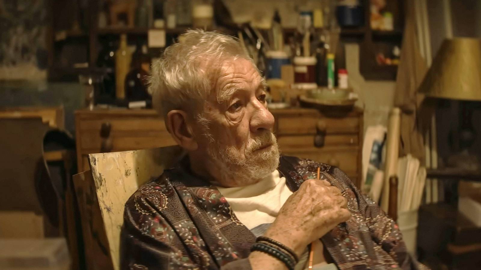 Ian McKellen seated in a chair inside a room filled with art supplies.