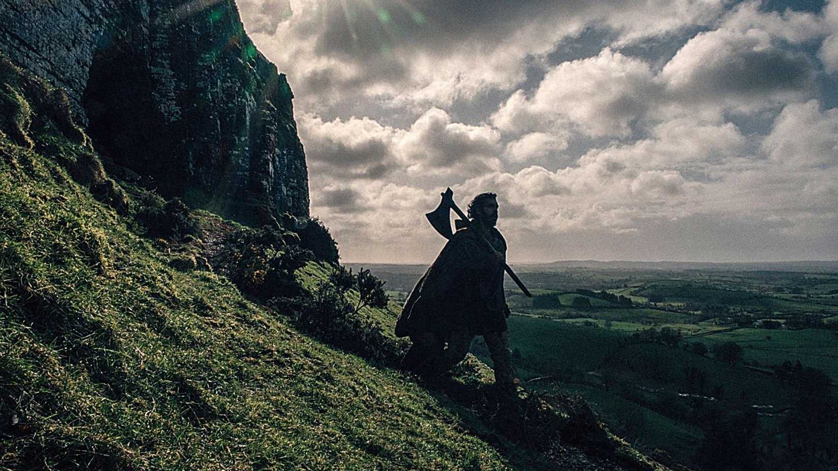 Gawain (Dev Patel) walking down a hill, carrying an axe in The Green Knight.