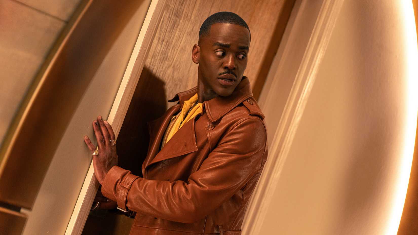 The Fifteenth Doctor entering through a doorway, wearing his brown trench coat 