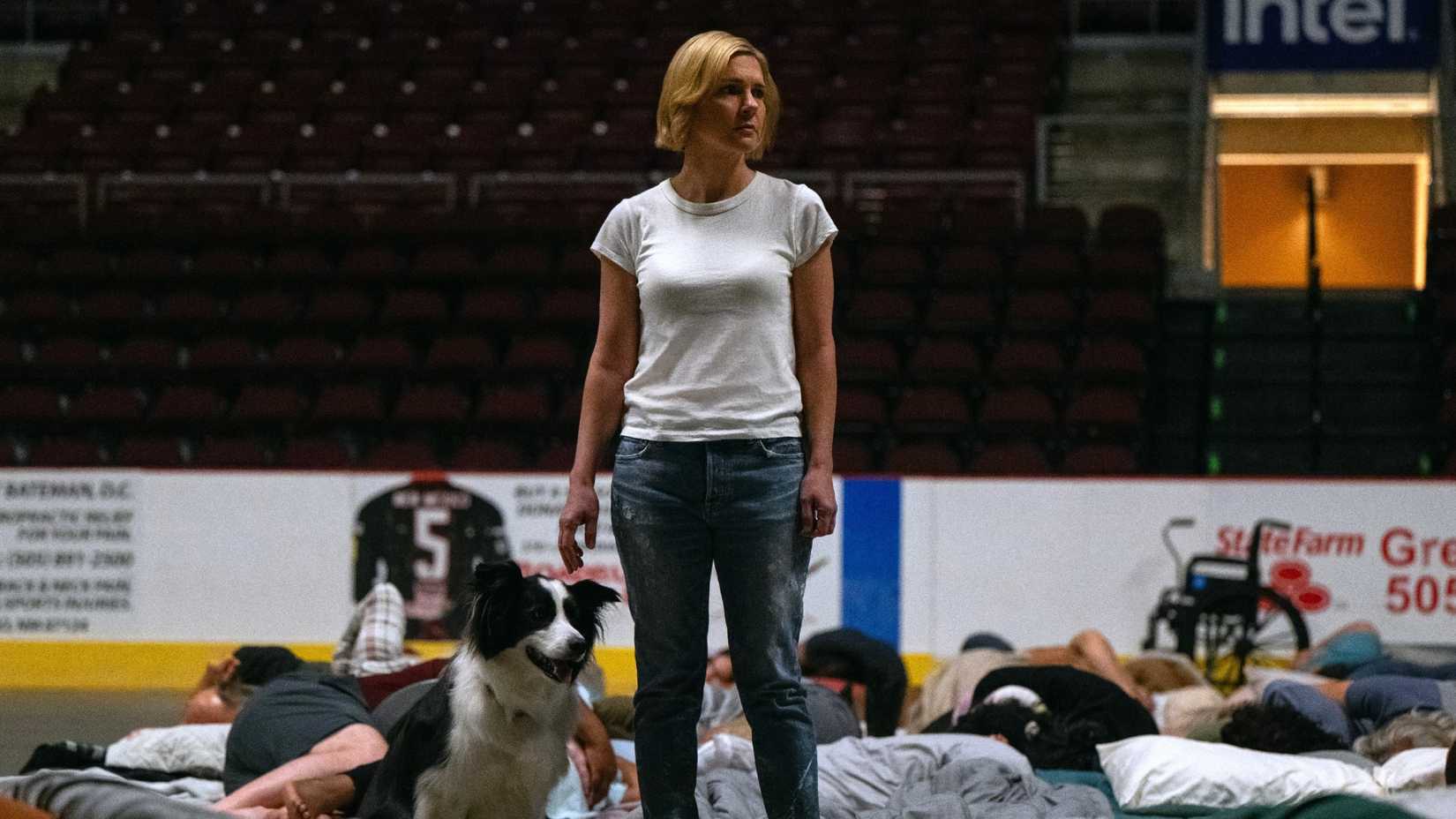 Rhea Seeborn's Carol stands in a stadium full of bodies with a dog from Pluribus