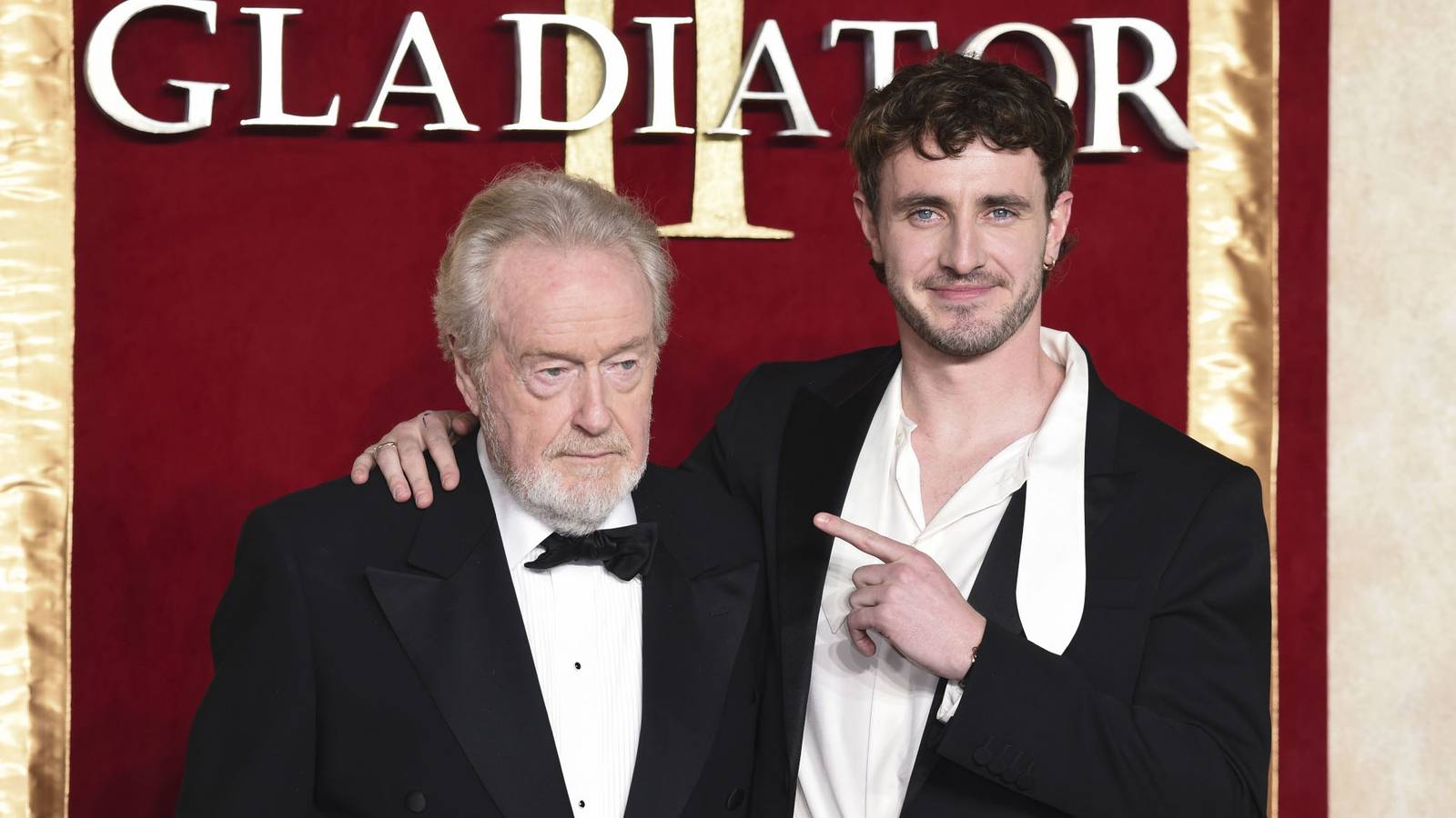 Paul Mescal smiles and points at scowling Ridley Scott while attending a red carpet event.