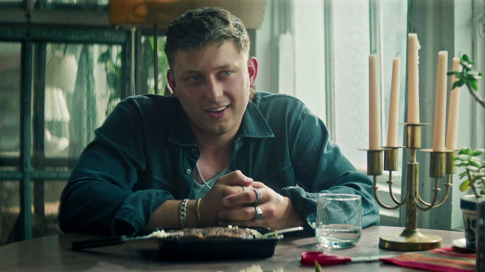 Clayton Angevine (Tom Francis) smiling at a table with his hands folded in You.