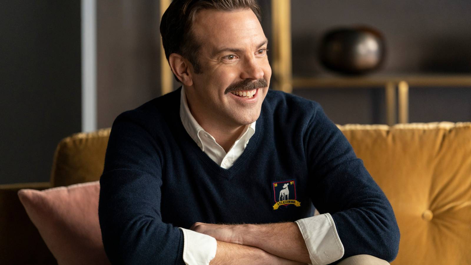 Ted Lasso (Jason Sudeikis) sitting on a couch and grinning in Ted Lasso season 2-1