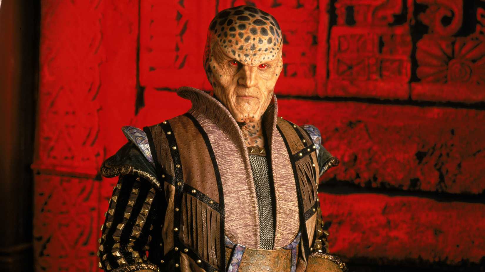 Andreas Katsulas‘ G’Kar standing in front of a red wall in Babylon 5