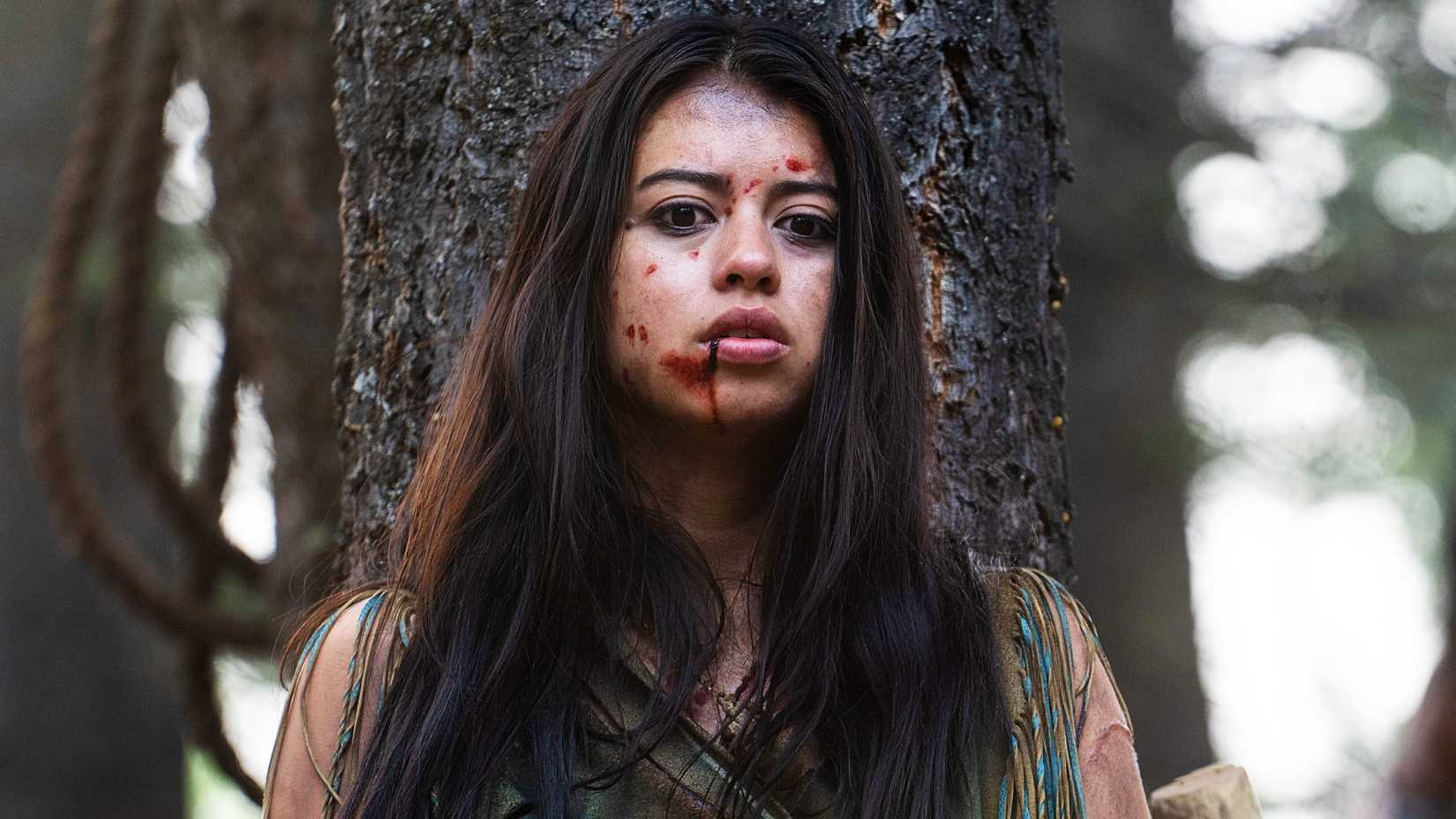 Amber Midthunder as Naru with blood on her face hiding behind a tree in Prey