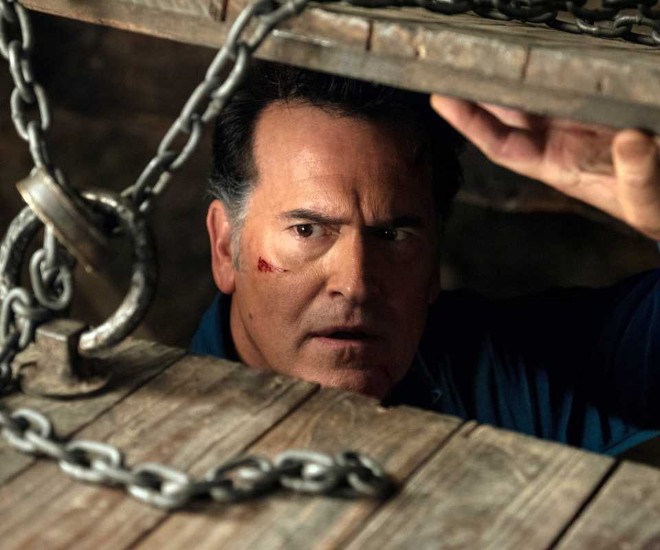Bruce Campbell's Ash looking up through the chained basement door suspiciously in Ash vs Evil Dead