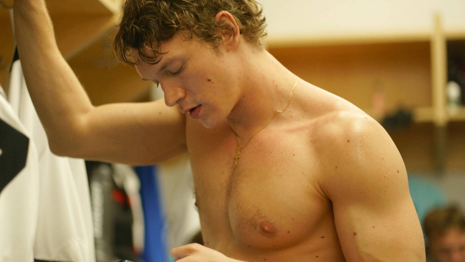 Connor Storrie as Ilya Rozanov checking his phone in the locker room shirtless in Heated Rivalry
