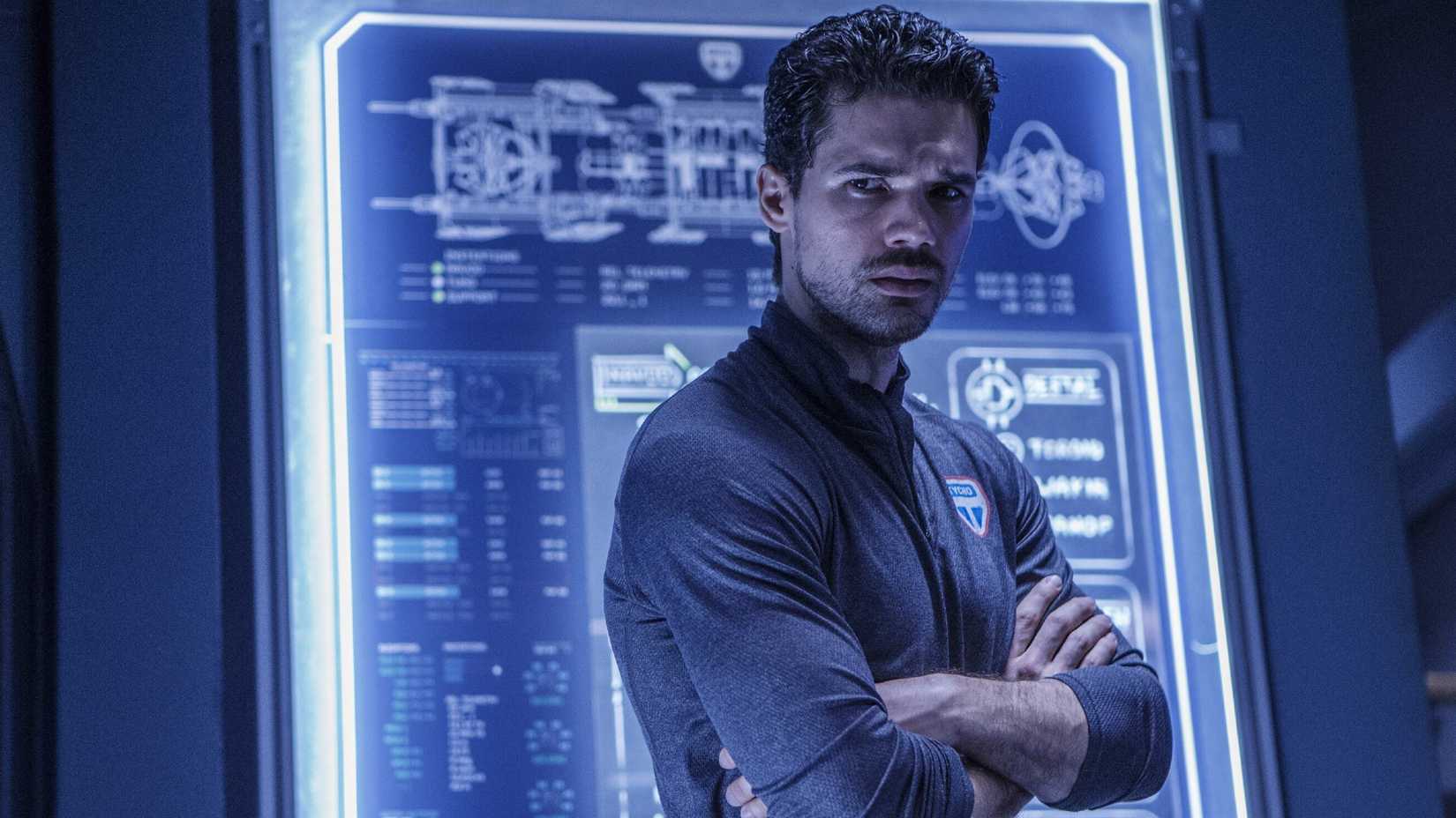 Jim Holden looks over his shoulder while standing in front of a glowing blue wall in The Expanse