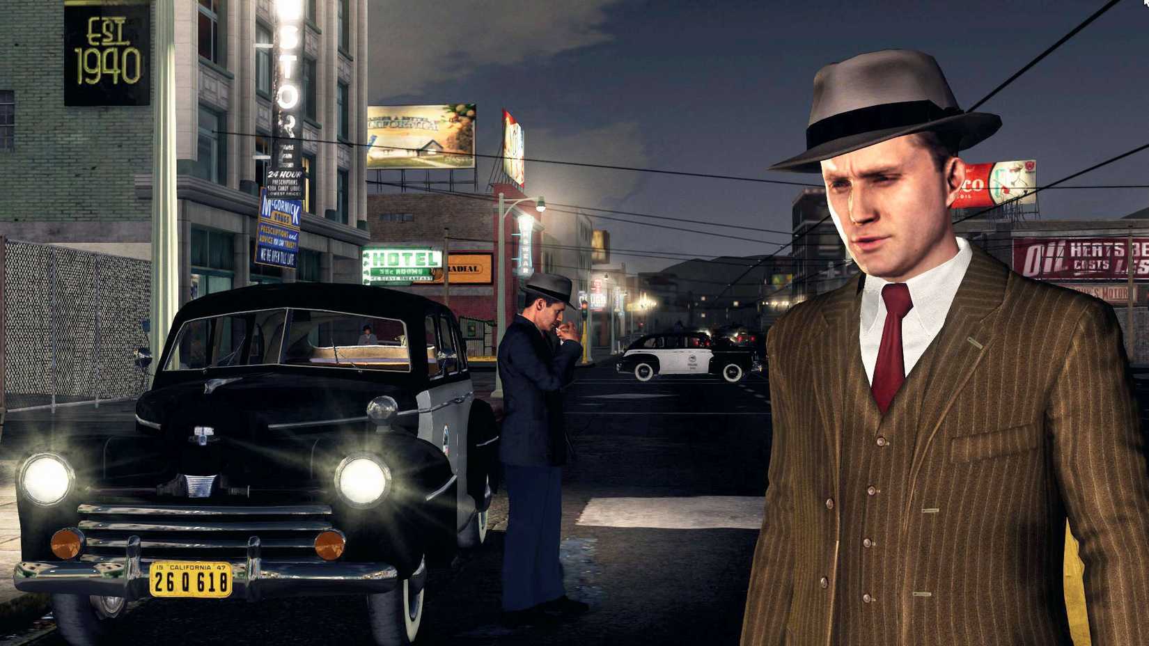 Cole standing in the foreground, with his partner next to a cop car behind.