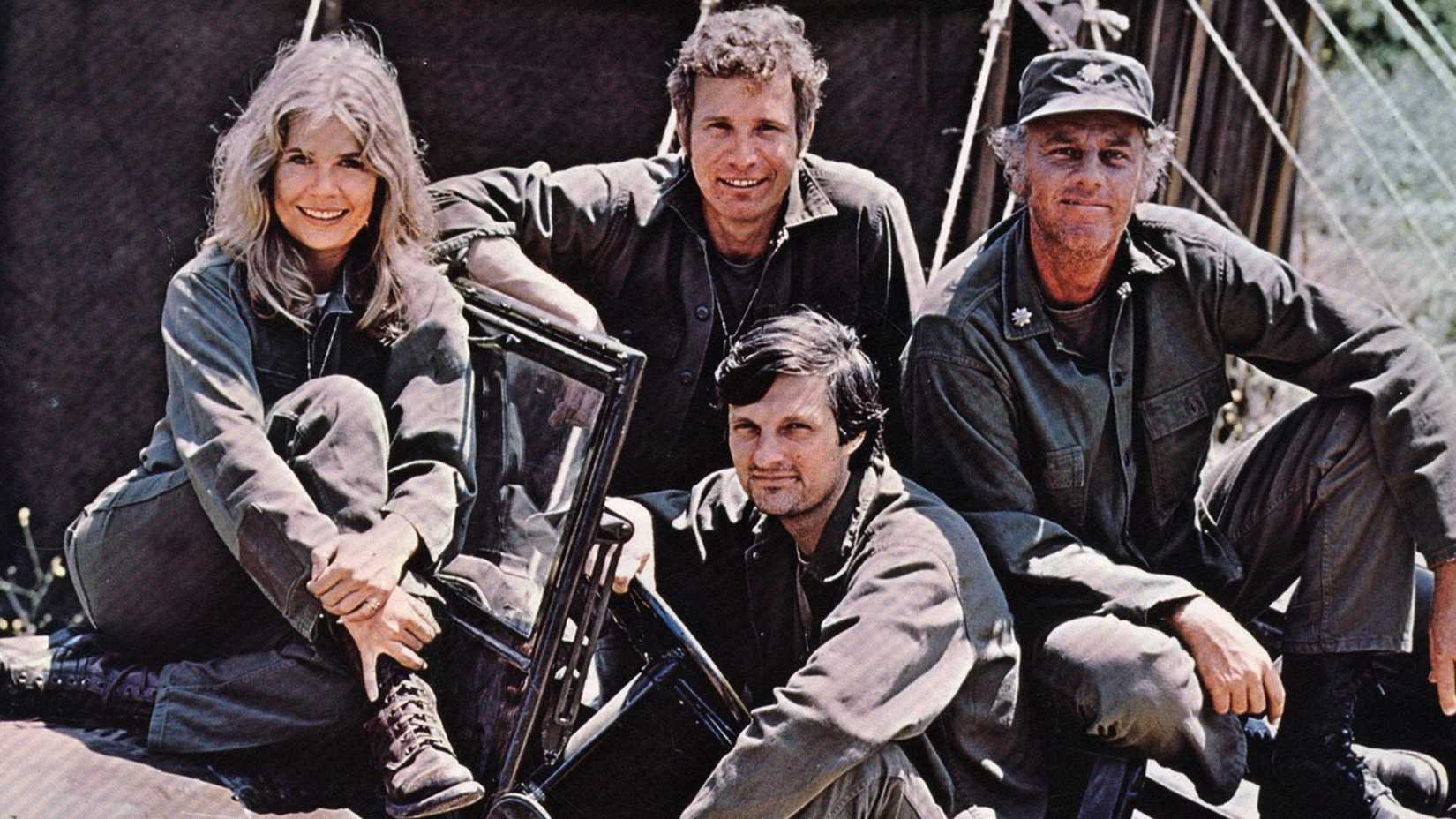 The cast of MASH wearing their fatigues and sitting in a Jeep, looking at the camera and smiling.