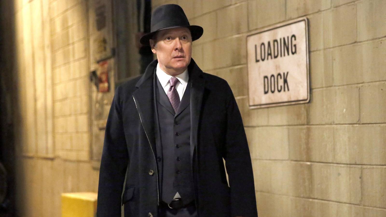 Still of James Spader as Raymond 'Red' Reddington standing in a suit and hat in front of gray brick wall in The Blacklist season 5 episode 15.