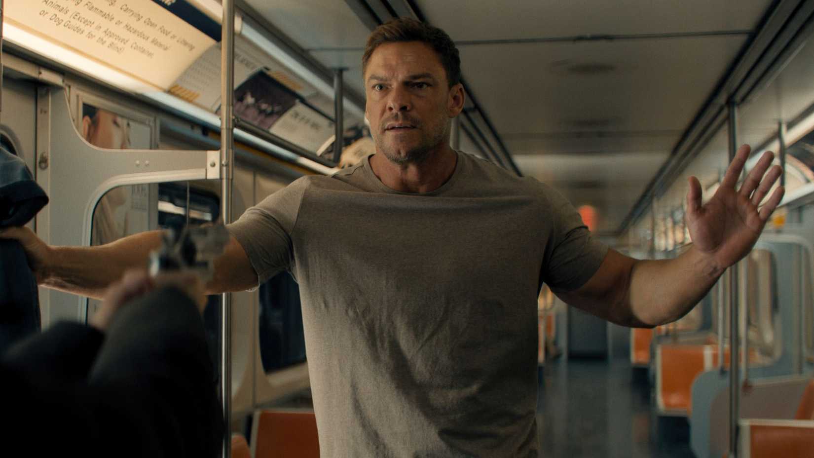 Alan Ritchson as Jack Reacher, looking frightened on a subway, with his hands held up
