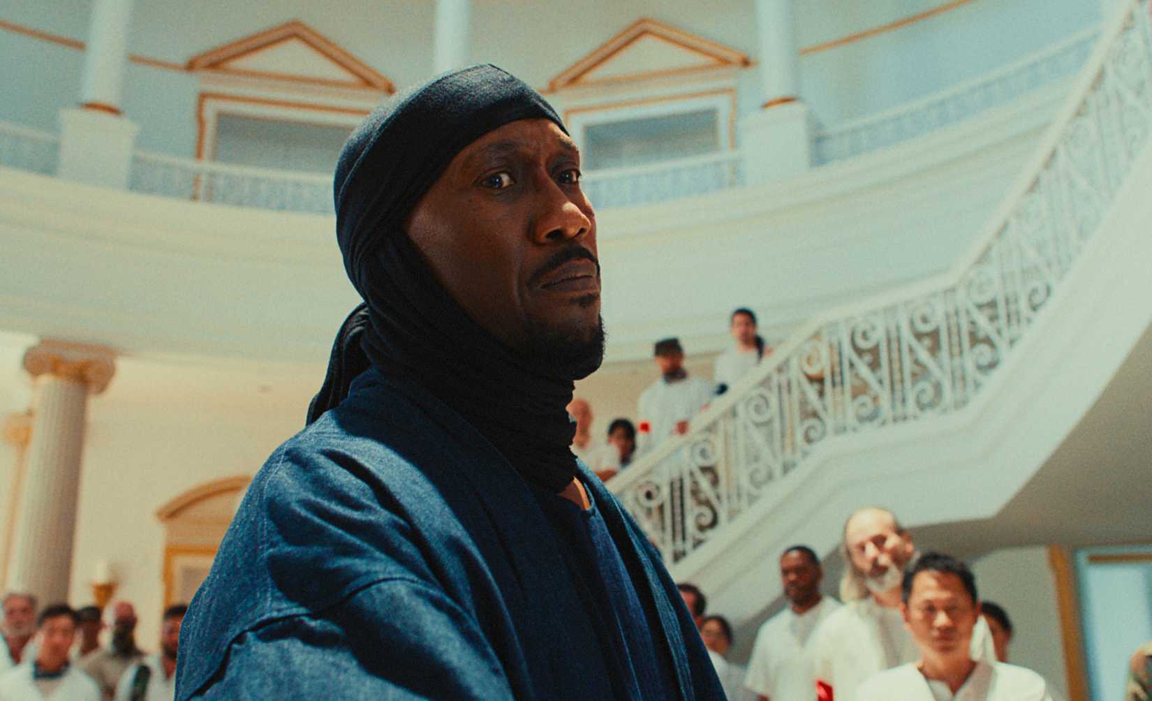 Mahershala Ali with an enigmatic look with a crowd of people wearing white behind him