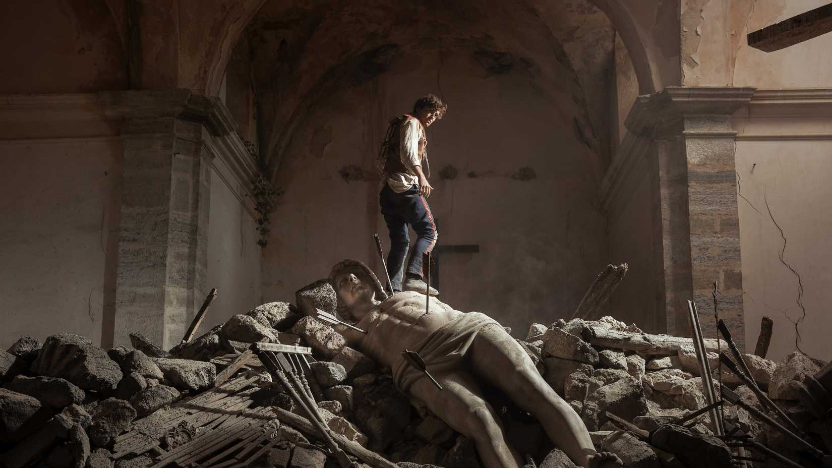 A man stands on top of ruins including an arrow-pierced statue in La Bola Negra