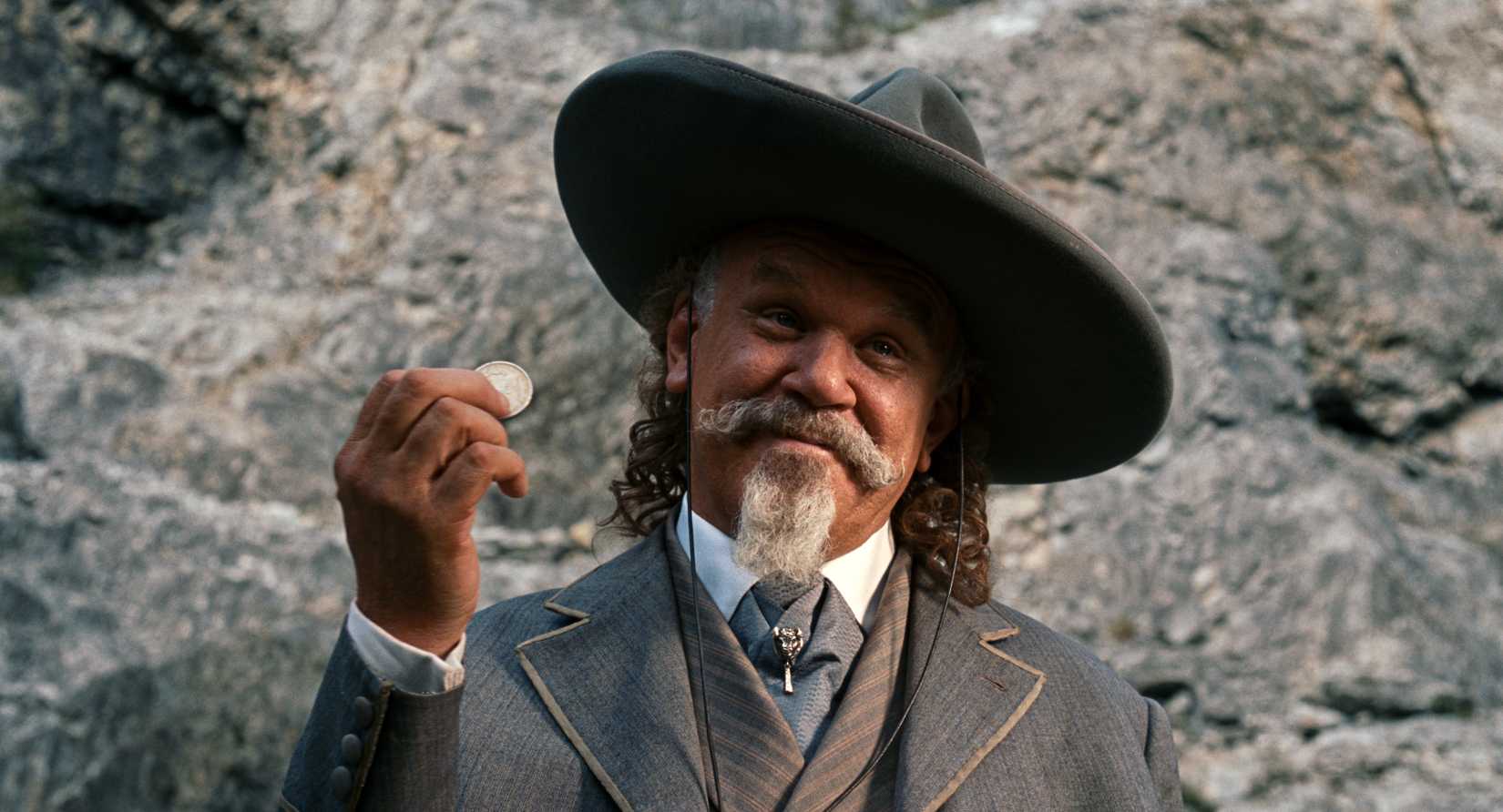 John C. Reilly holds a coin up and grins at someone off camera in  Heads or Tails 