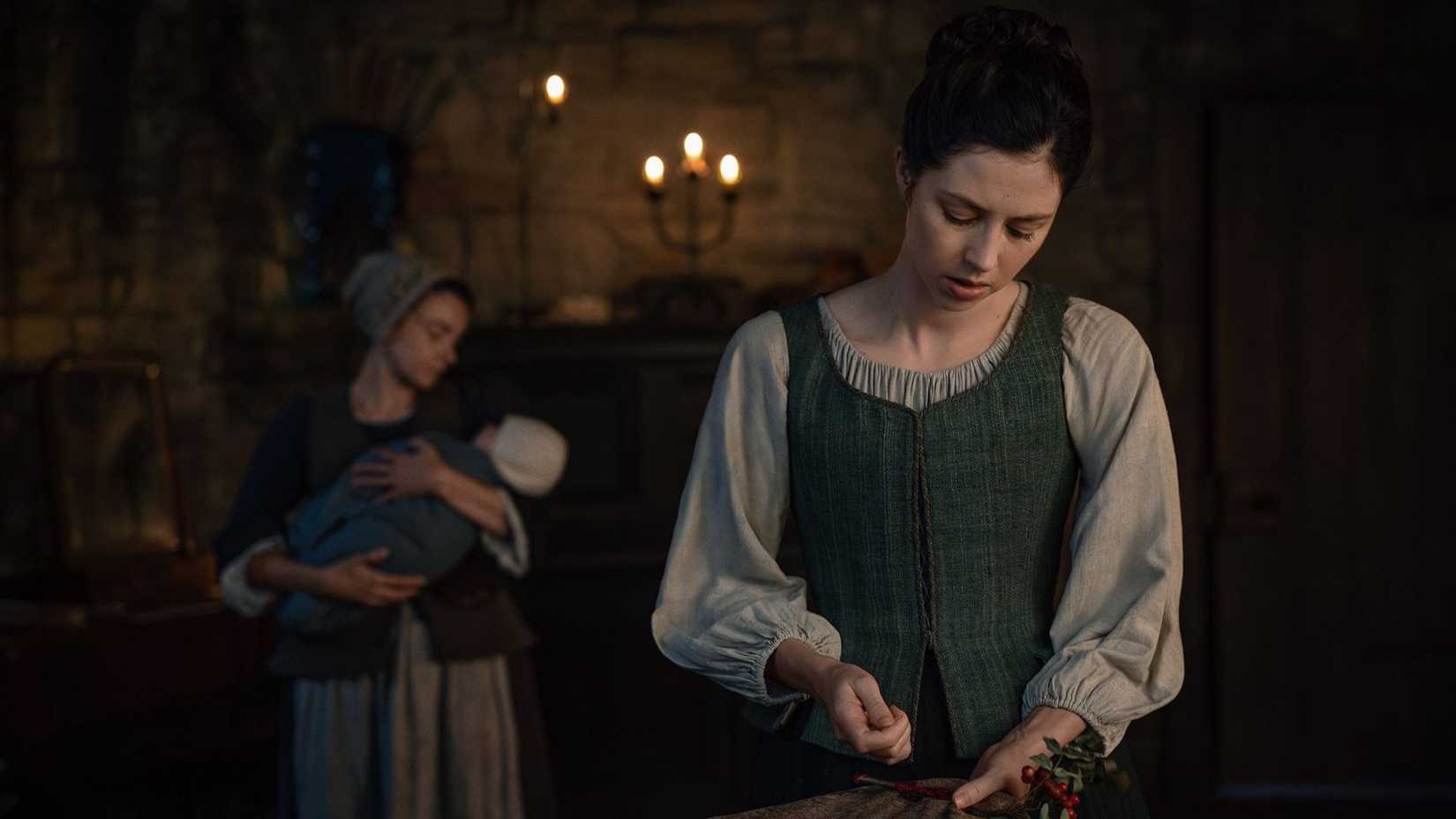 Julia cooking while Davina is behind her with the baby in Outlander: Blood of My Blood