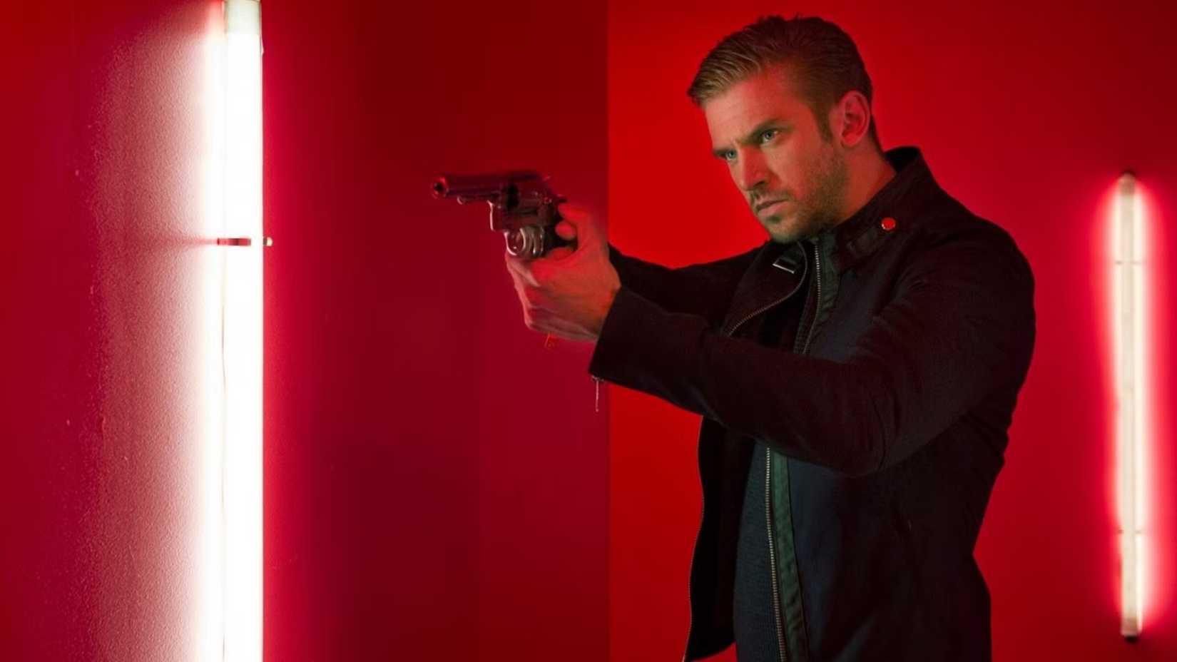 Dan Stevens aiming a revolver against a red background in The Guest