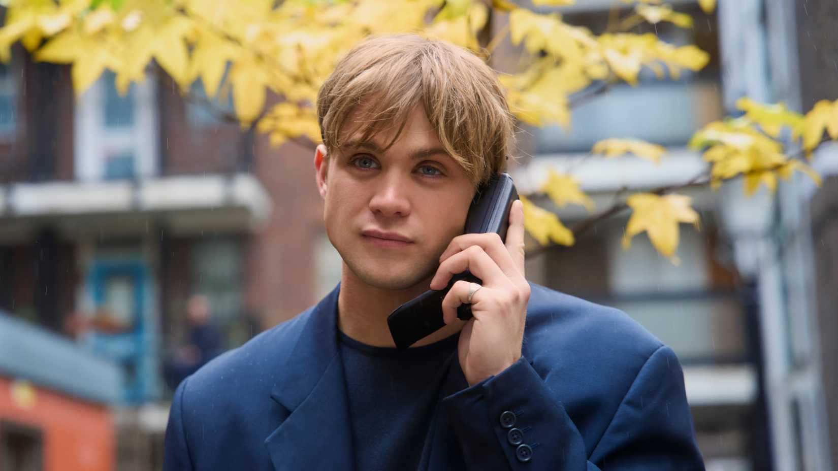 Dex smiles while talking on the phone and standing in front of a tree with yellow leaves in One Day