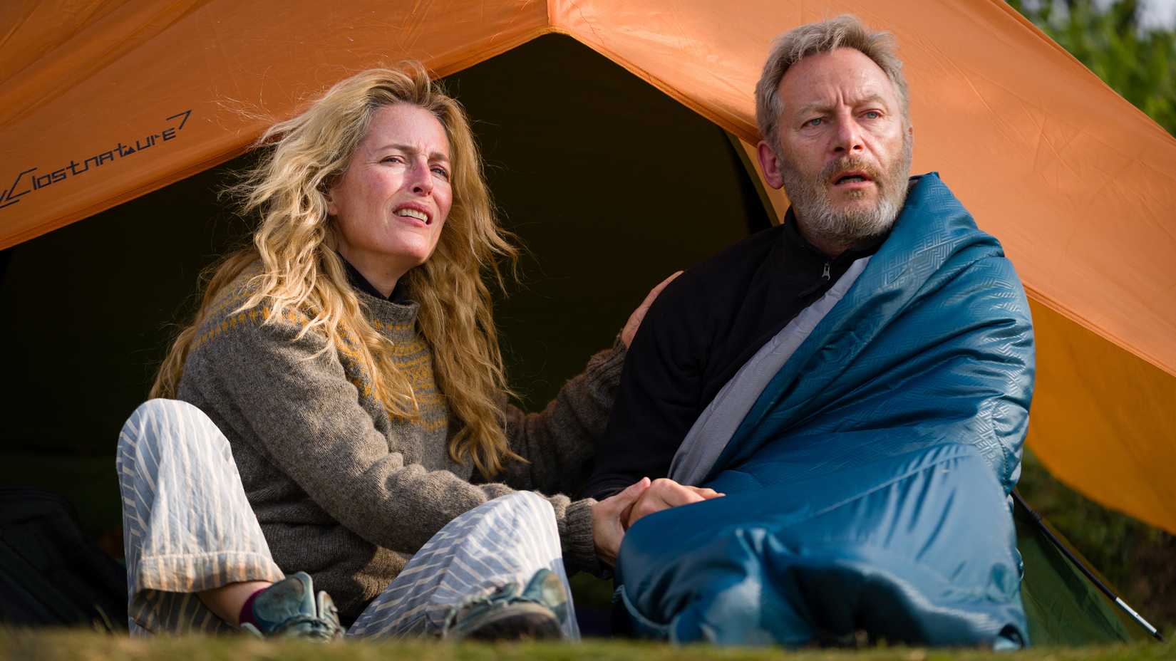 Gillian Anderson's Raynor and Jason Isaacs' Moth looking uncertainly at something while sitting outside of their tent in The Salt Path