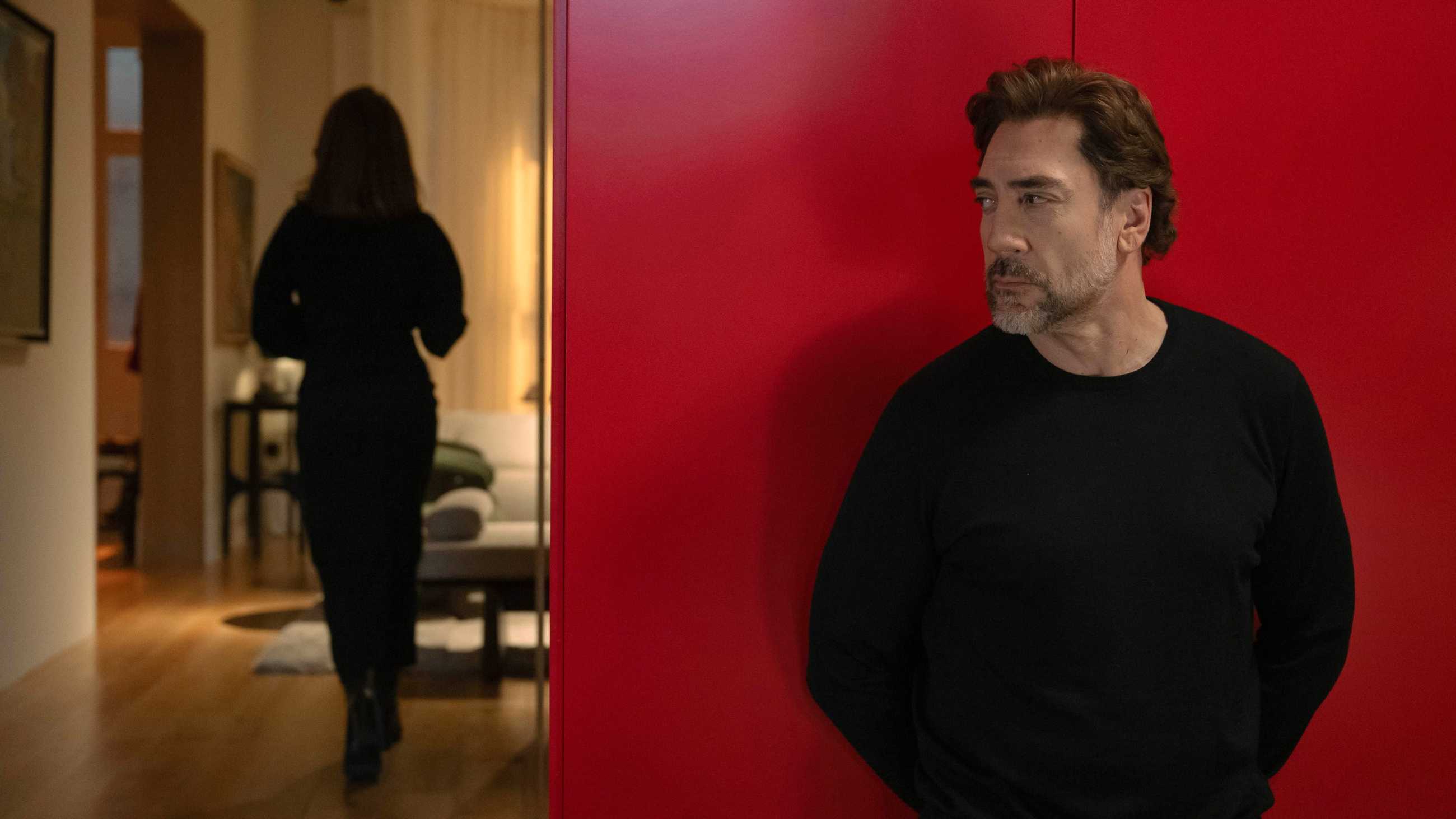 Javier Bardem standing against a red closet as a woman walks down a hallway in Bunker